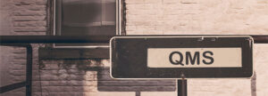 A "One Way" directional sign labeled 'QMS' on a brick wall, serving as a visual metaphor for qmsWrapper’s integrated approach that mirrors Project, Quality, and Document Management into a single compliance DNA.