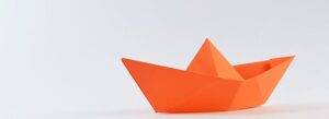 An orange origami paper boat on a white background, symbolizing the "golden middle path" of QMS documentation that balances regulatory compliance with operational efficiency.